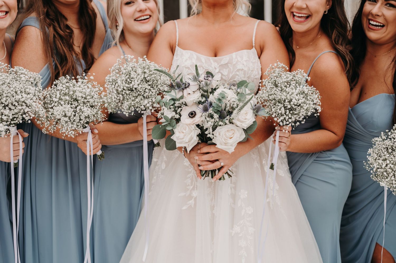 Bridesmaids in dusty blue dresses holding baby's breath bouquets next to bride in white.