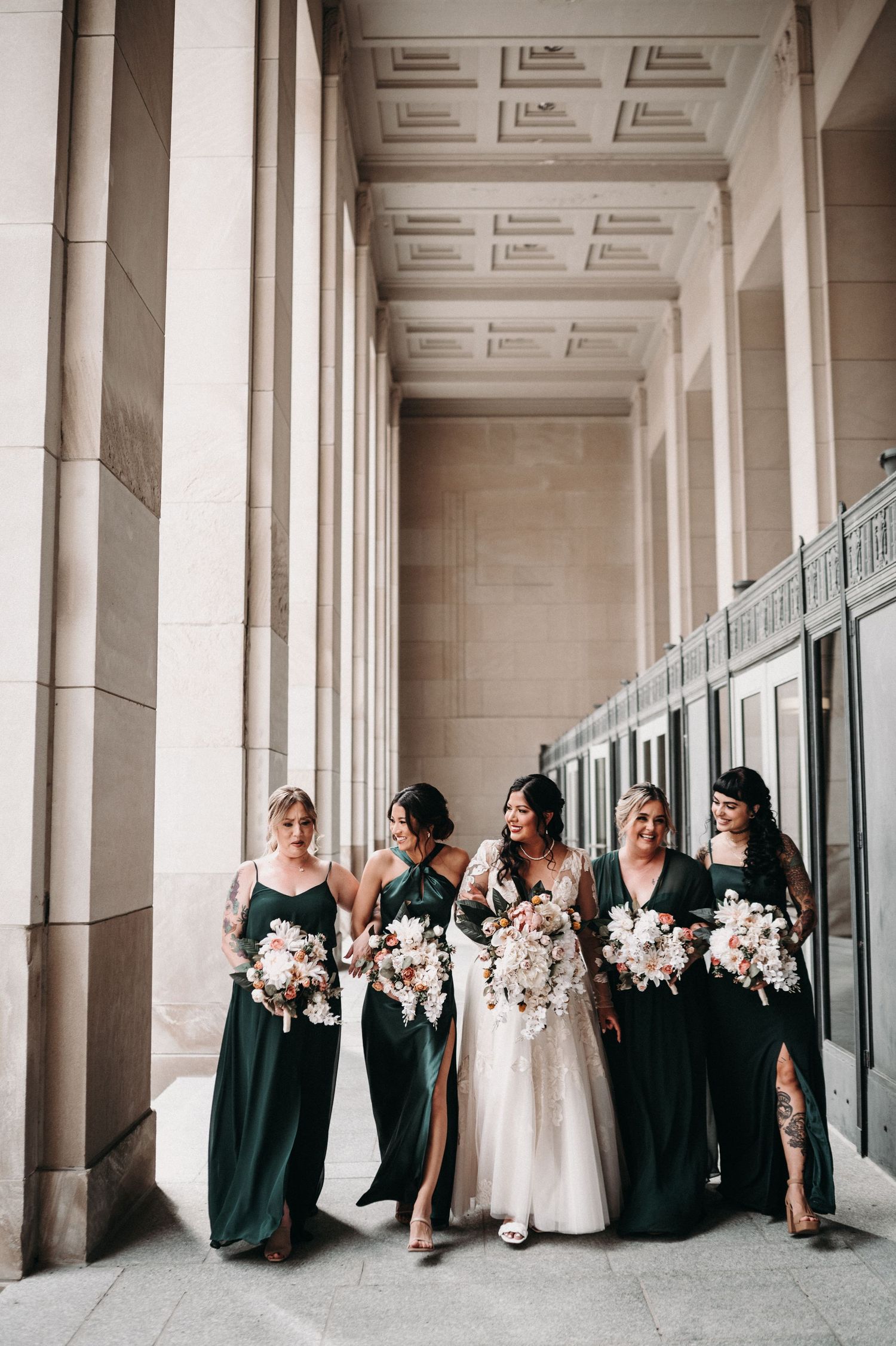 Bridesmaids in dark green gowns laugh with bride while holding pink and white bouquets.