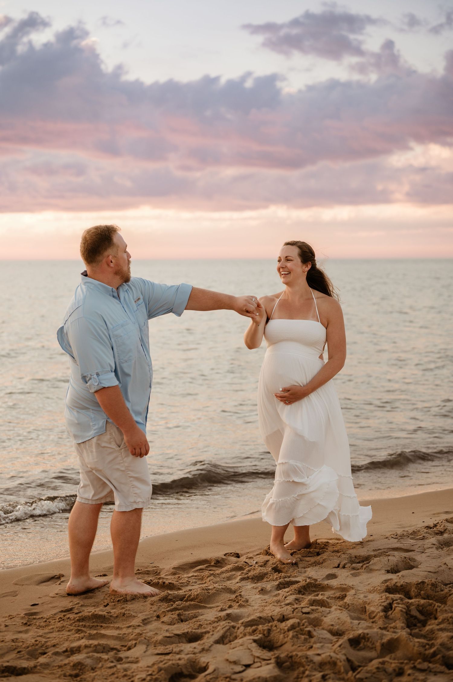 A couple shares a romantic moment on a beach at sunset with pink and purple clouds reflecting on the ocean.