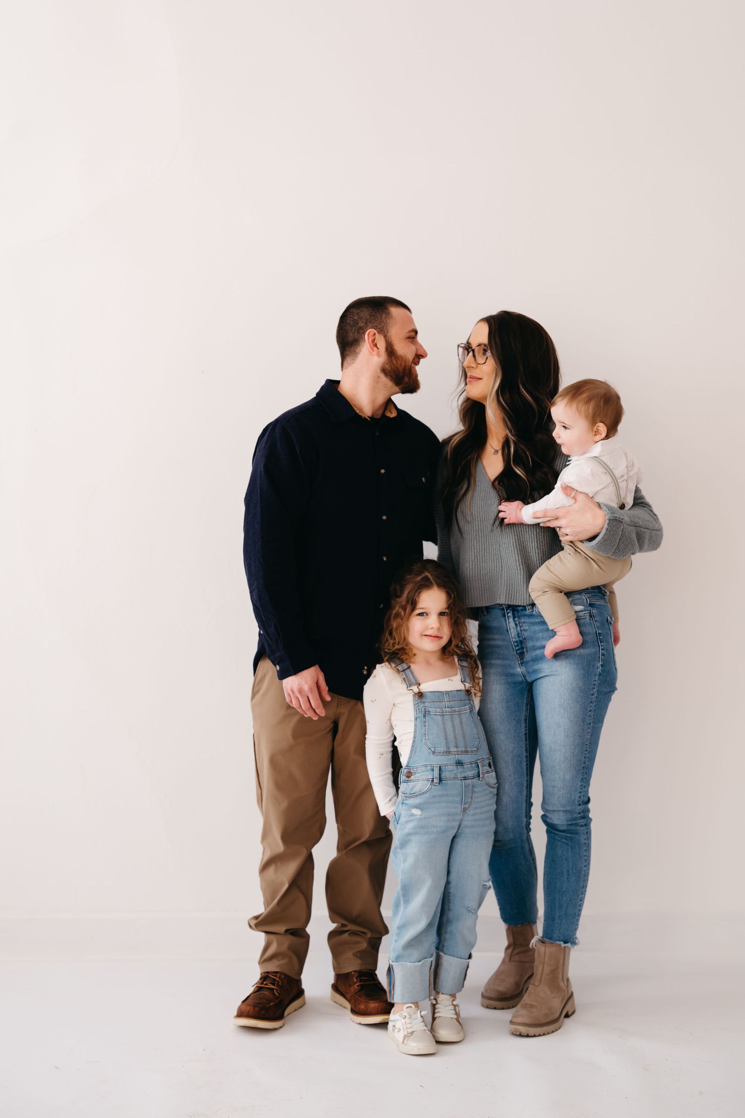 A family of four poses together against a white background wearing casual clothing in a minimalist style portrait.