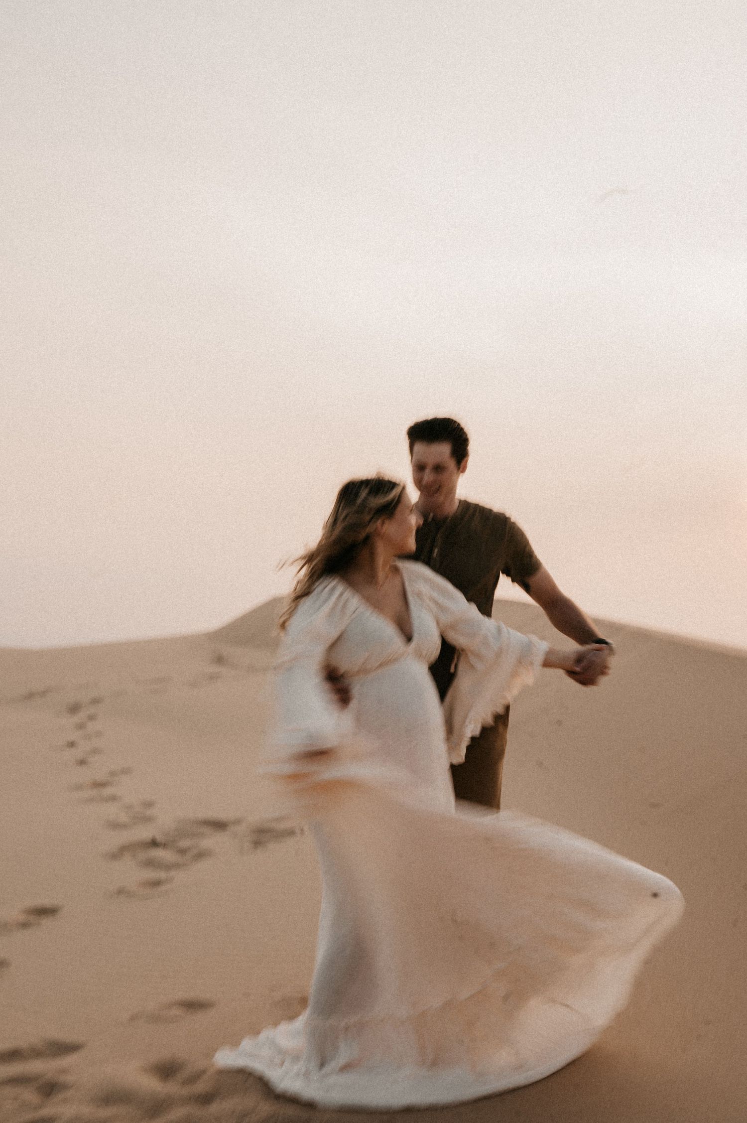 A couple dances joyfully on desert sand dunes during a romantic sunset photoshoot.