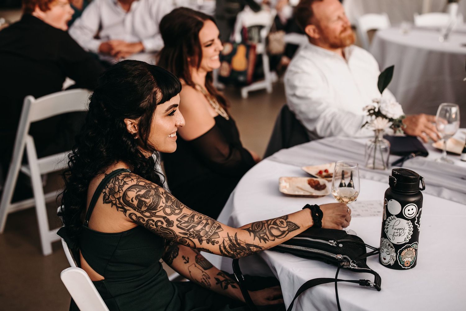 Wedding guests sit at a table with one person displaying intricate arm tattoos during a reception celebration.