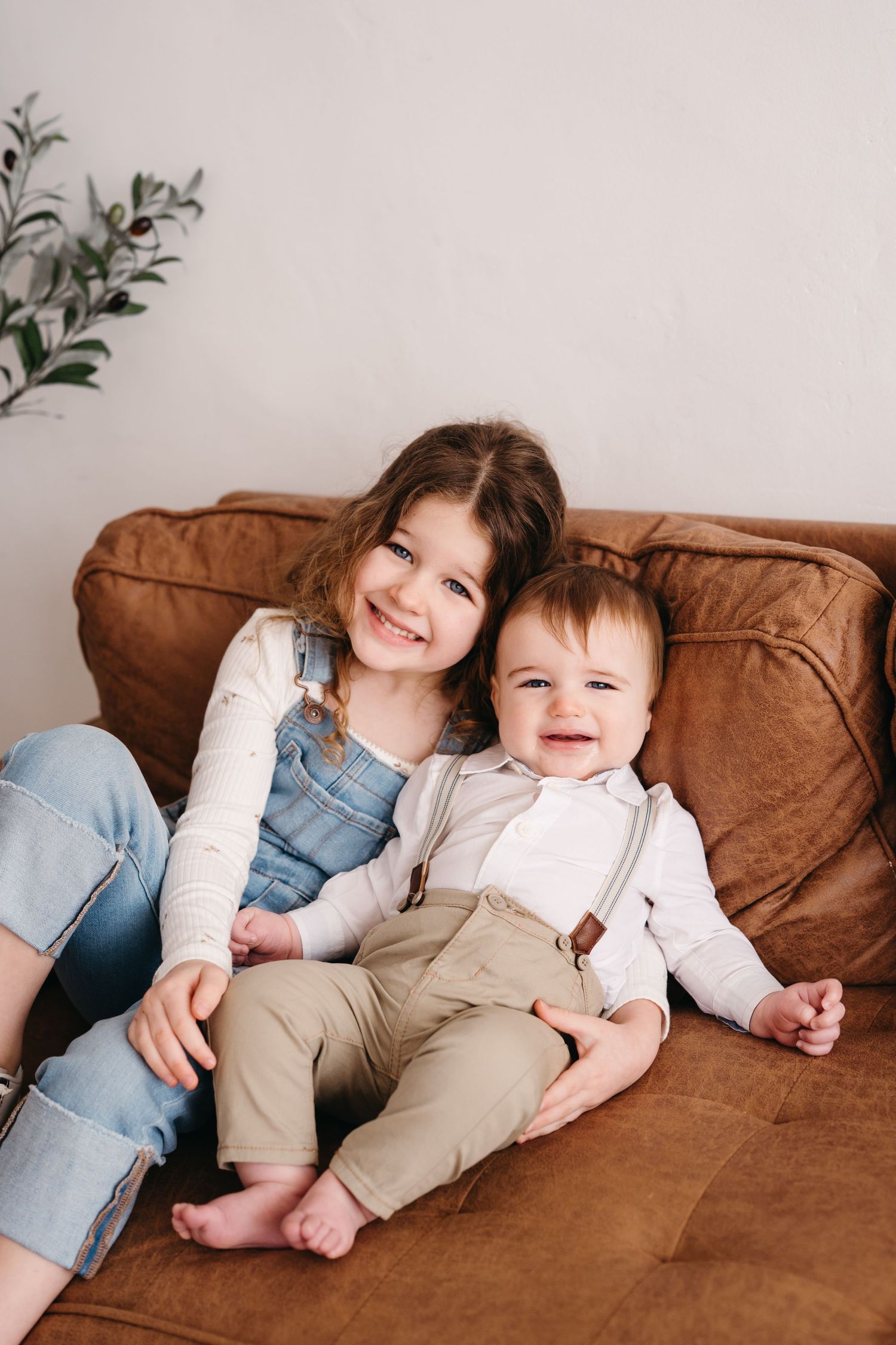 Two children sit together smiling on a brown leather couch while wearing casual clothes.