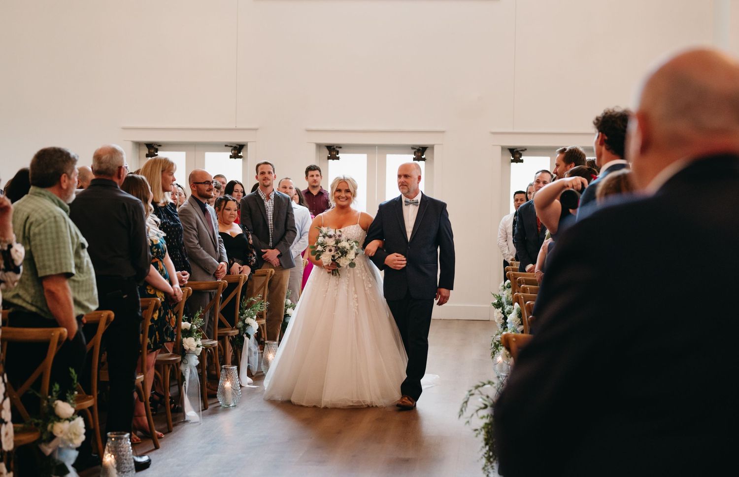 A wedding ceremony processional down the aisle in a bright indoor venue with guests seated on wooden chairs.