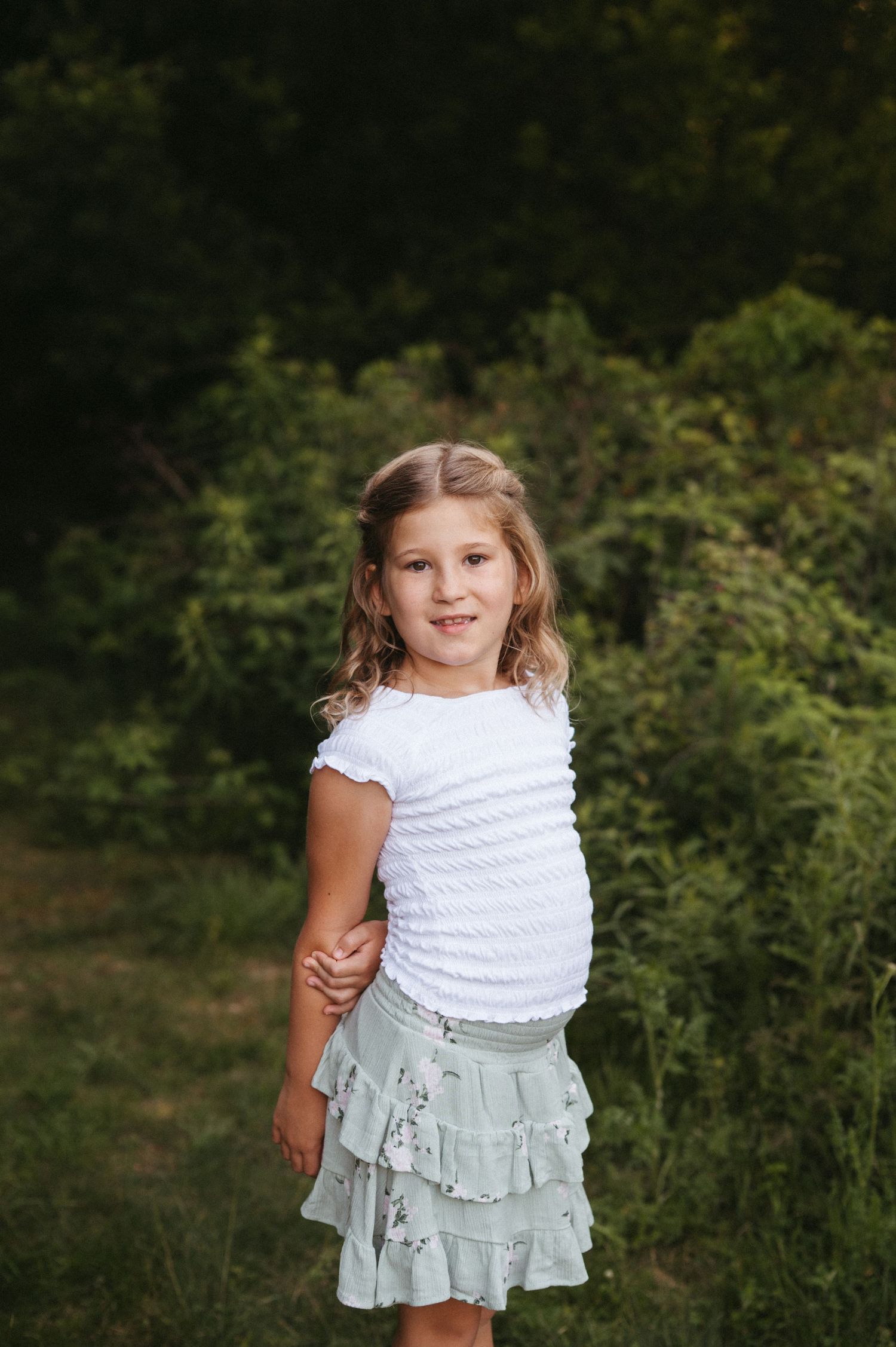 Young child in a white ruffled top and mint green skirt poses outdoors against a green natural background.