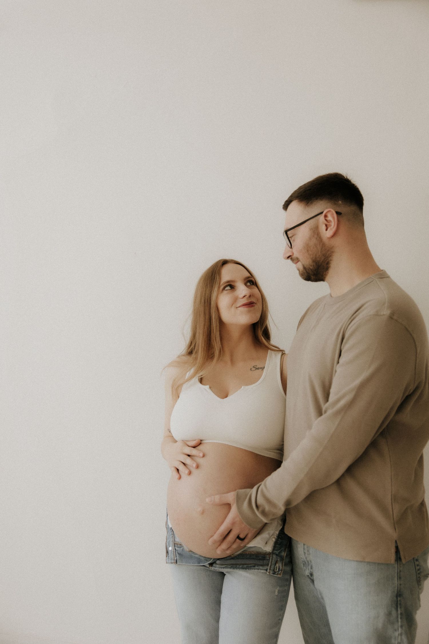 Intimate maternity photoshoot showing couple in casual attire against white wall.