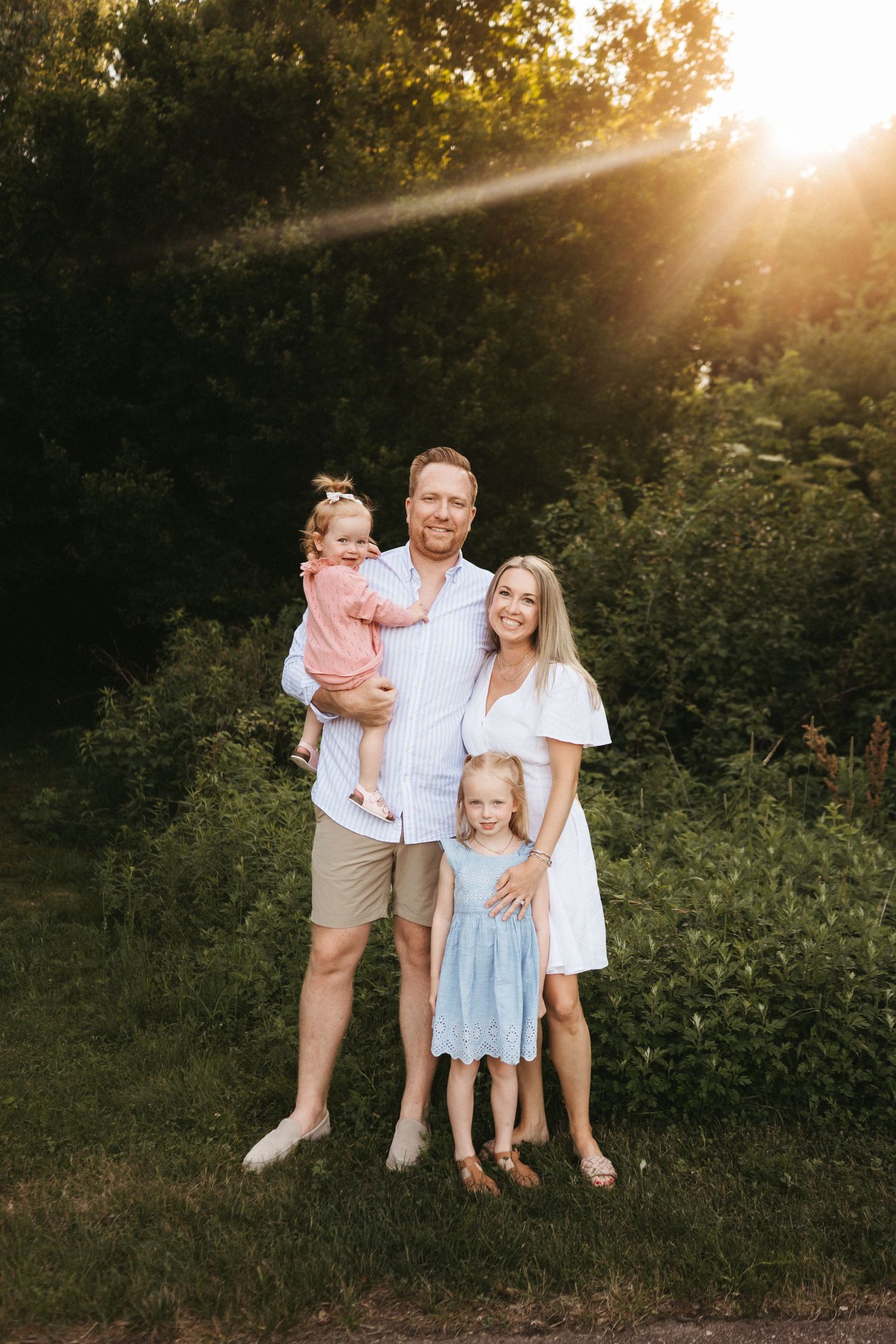 A family of four poses together outdoors at sunset with golden sunlight streaming through trees in the background.