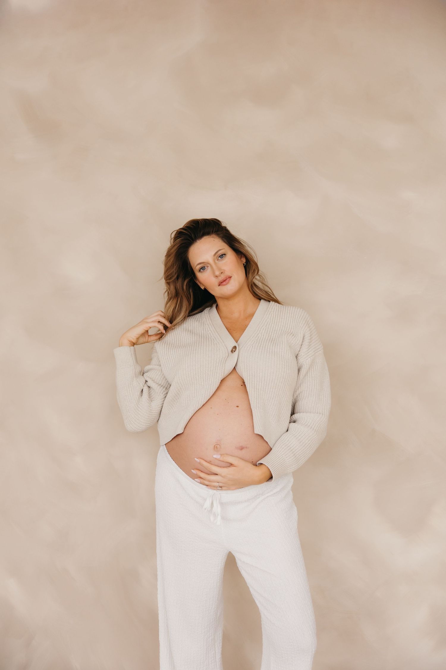 Pregnant woman in open beige cardigan and white pants holding her bare baby bump against beige wall.