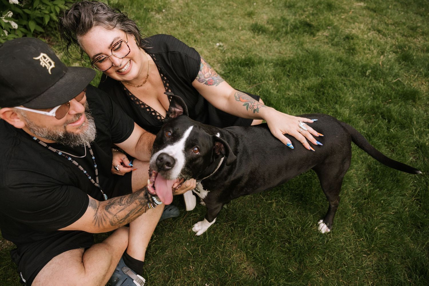 Couple in black clothing sitting on grass smiling with their black and white pit bull dog.