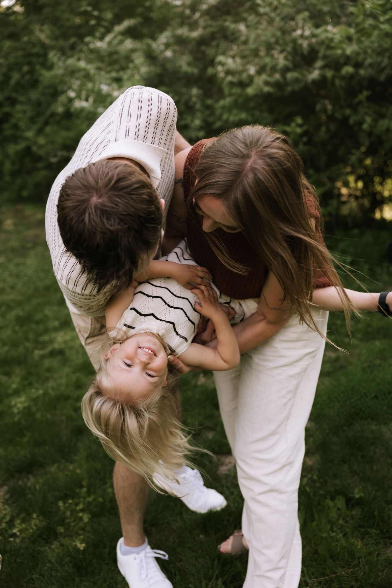 Parents playfully flip their laughing toddler girl upside down in a lush green backyard.