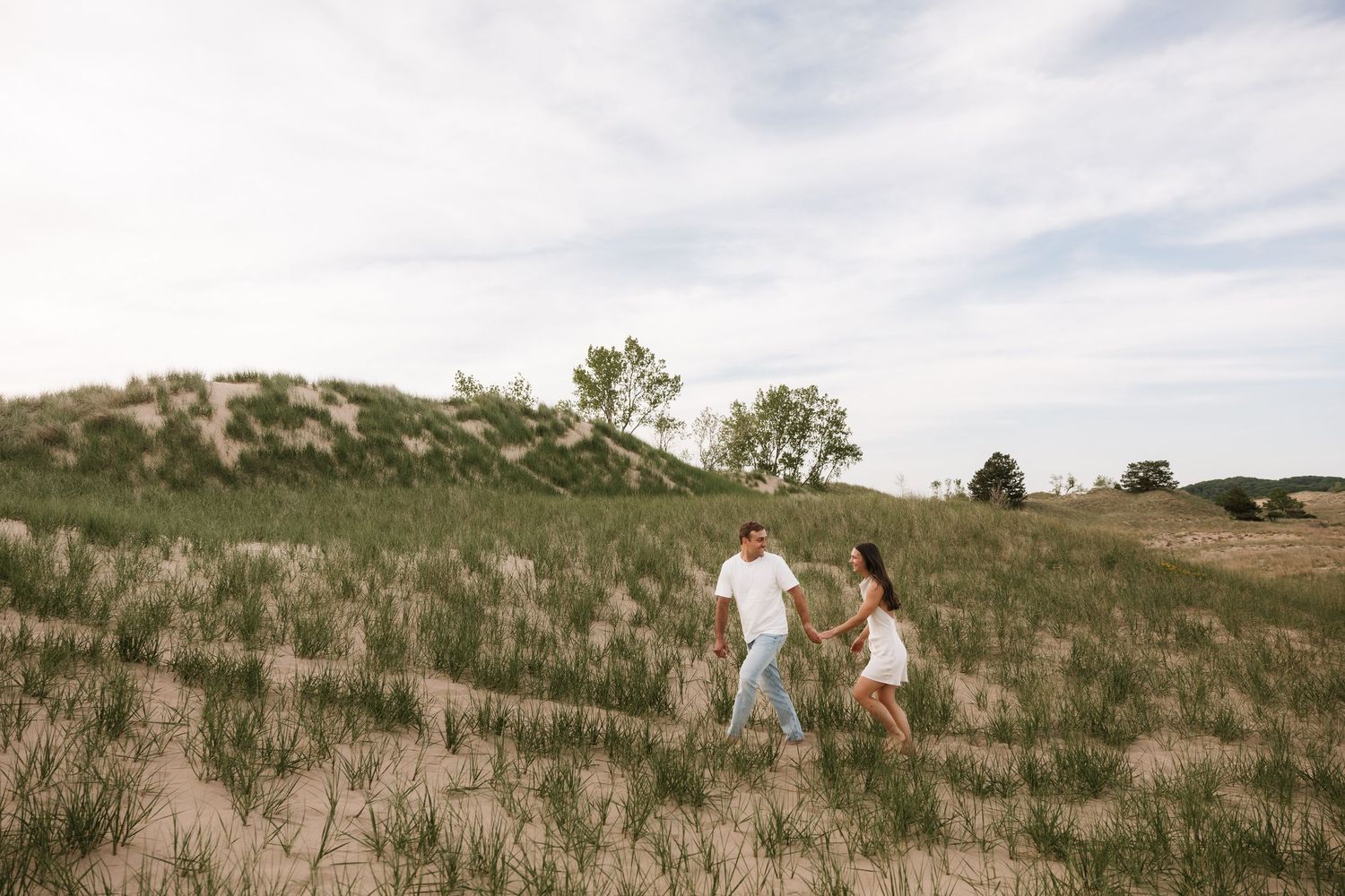 Couple walking hand in hand through sandy grass dunes under a cloudy sky in a romantic outdoor engagement session.