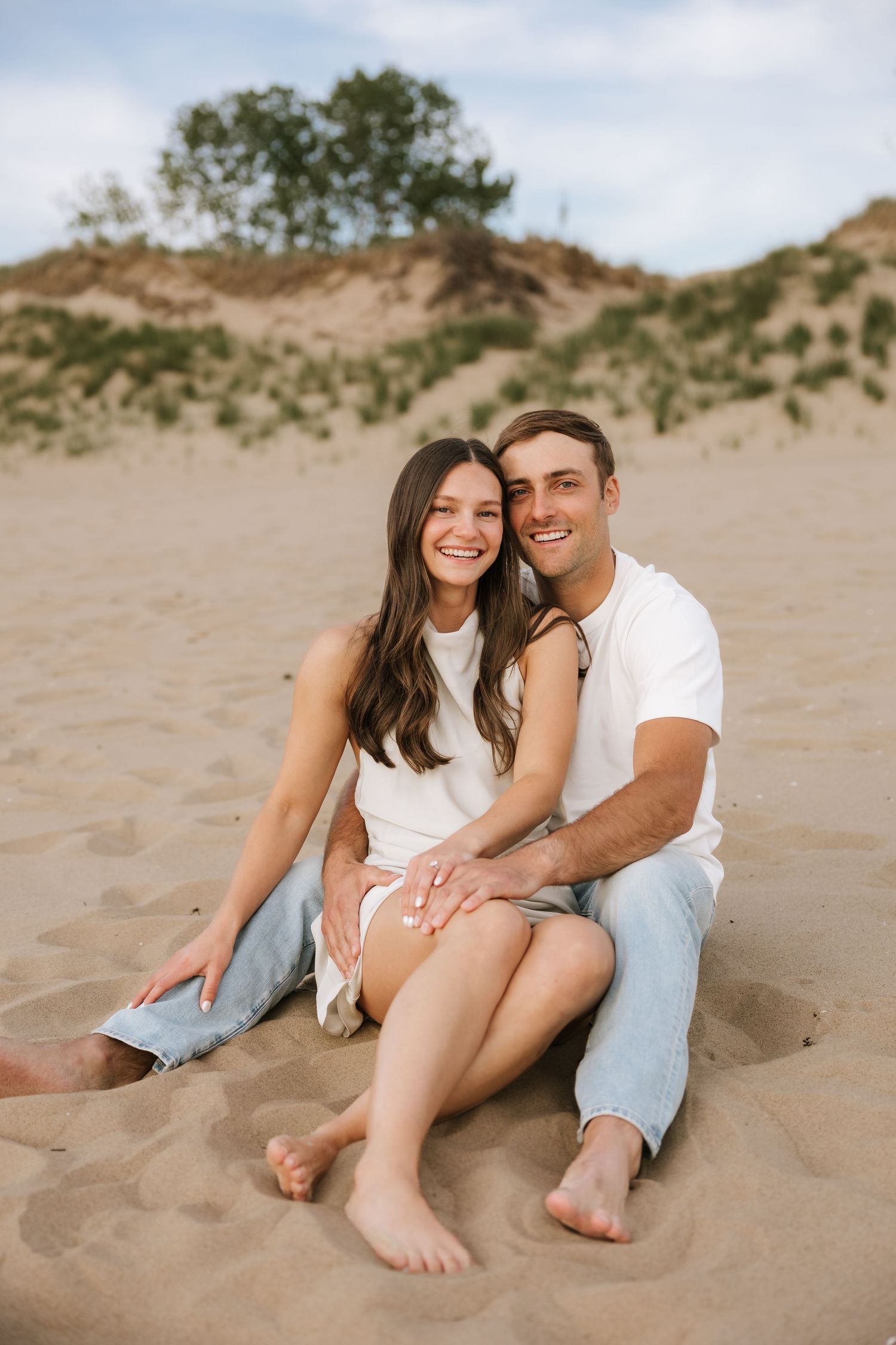 Smiling couple sitting together on sandy dunes, woman in white dress leaning against man in white tee.