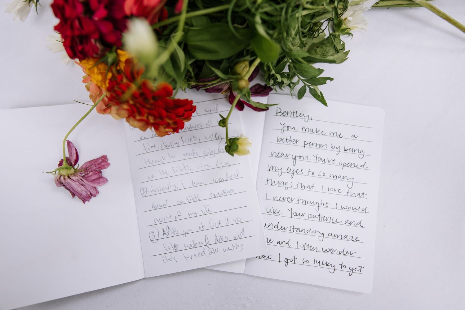 Handwritten note card displayed beside vibrant red and orange flowers with green stems on a white surface.