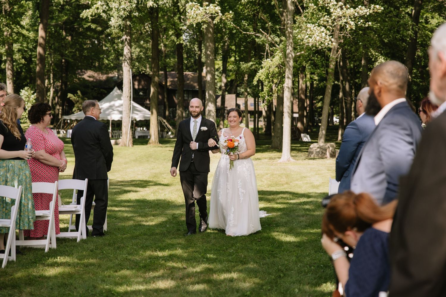 Wedding guests gather under white tents and trees during an outdoor summer celebration.