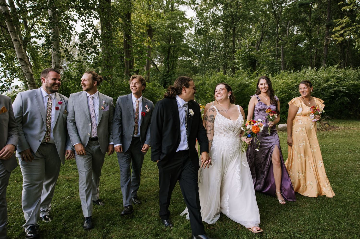 Wedding party in formal attire walking and laughing together on a green lawn.
