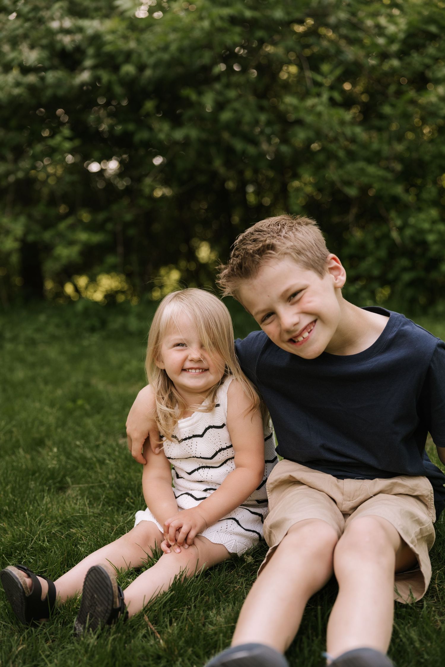 Two smiling siblings, a young boy and toddler girl, sit together on green grass in a sunny backyard.