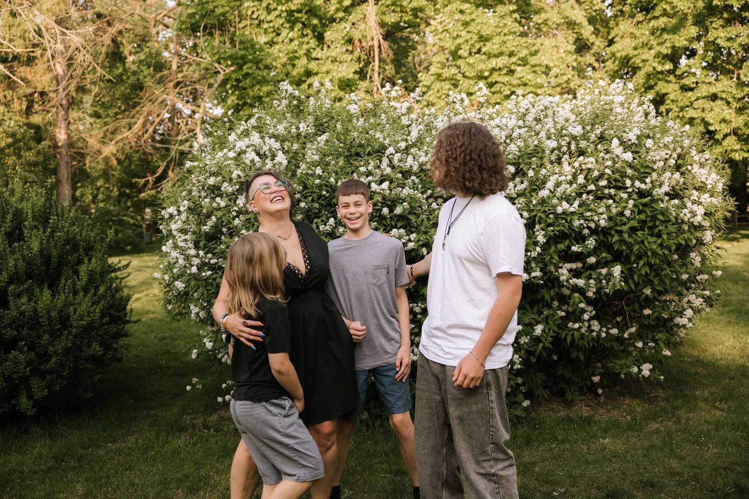 Group of four people laughing and embracing outdoors near white flowering bushes in summer.