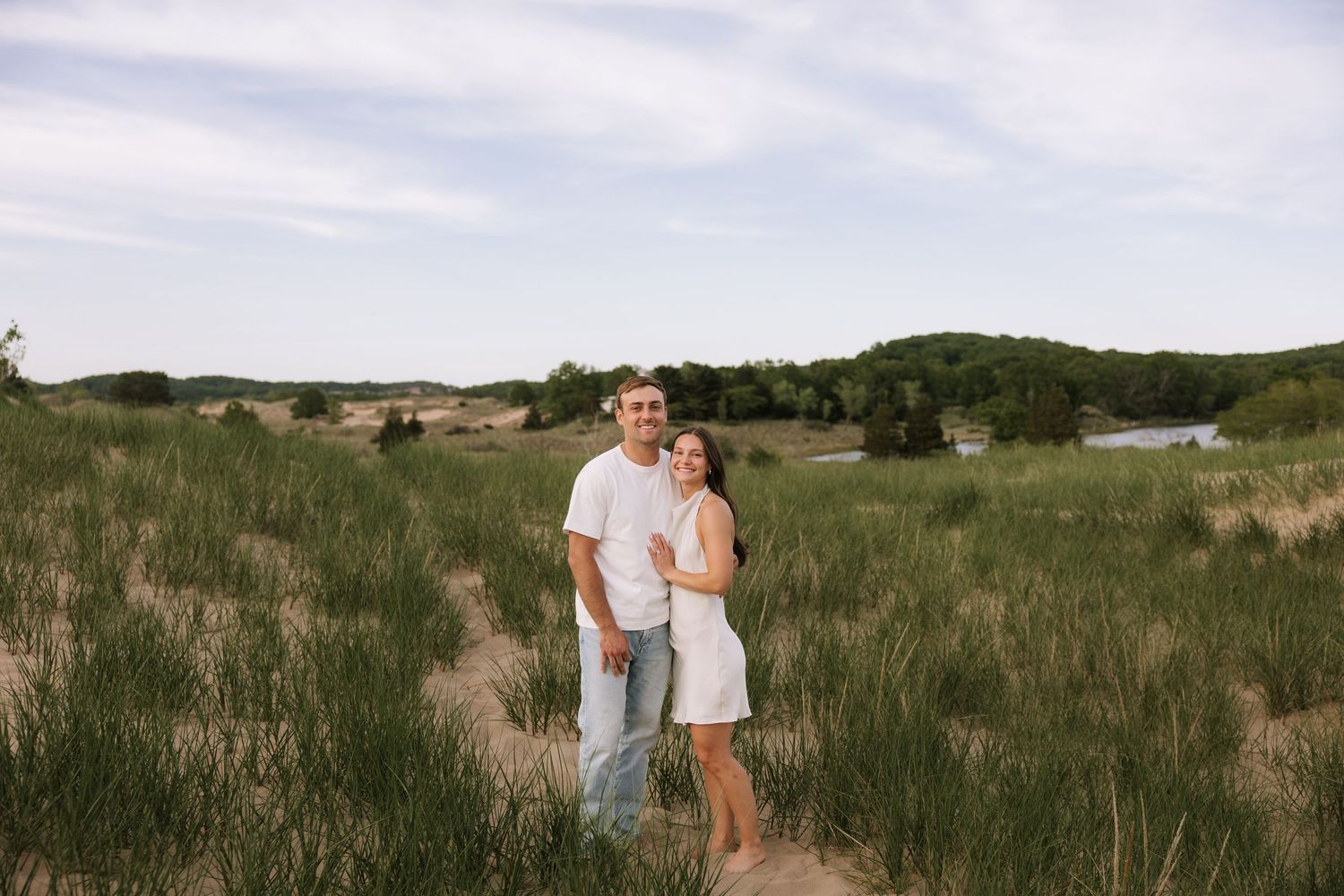 Couple smiling together in a lush green meadow with sandy dunes and a serene lake in the background.