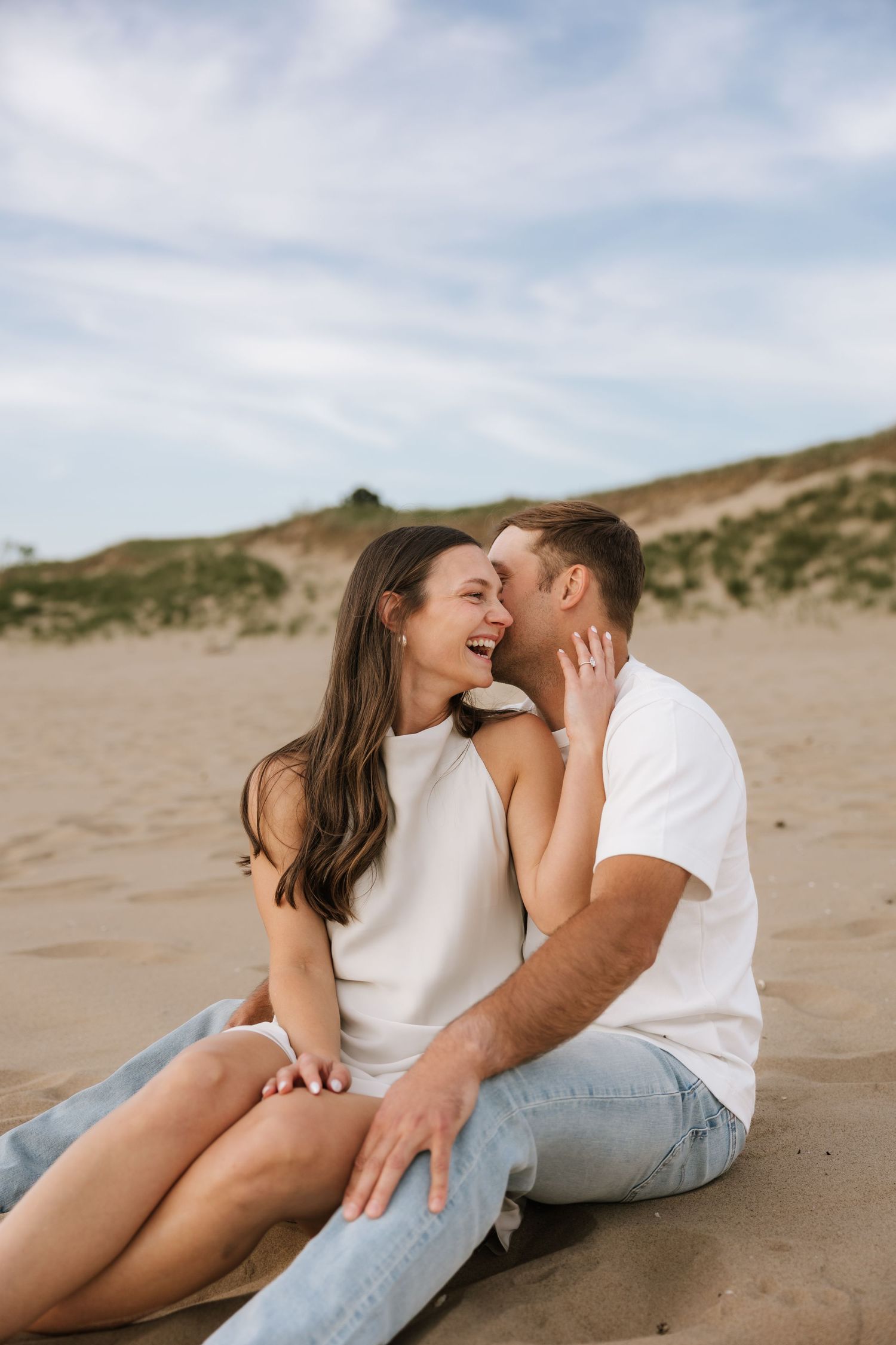 Joyful couple sitting close together on sandy beach dunes, laughing and sharing a moment.