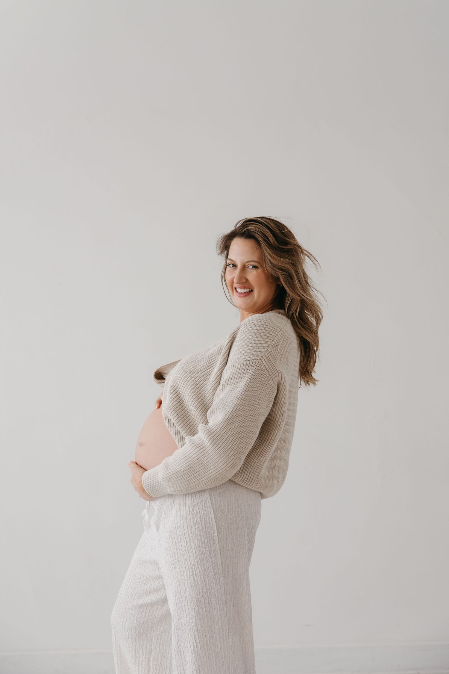 Smiling pregnant woman in cream ribbed sweater and pants cradling baby bump against white background.