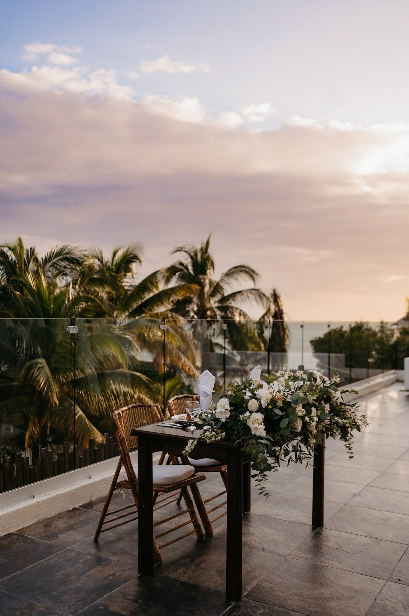 Sunset wedding reception setup with floral arrangements and palm trees in tropical setting.