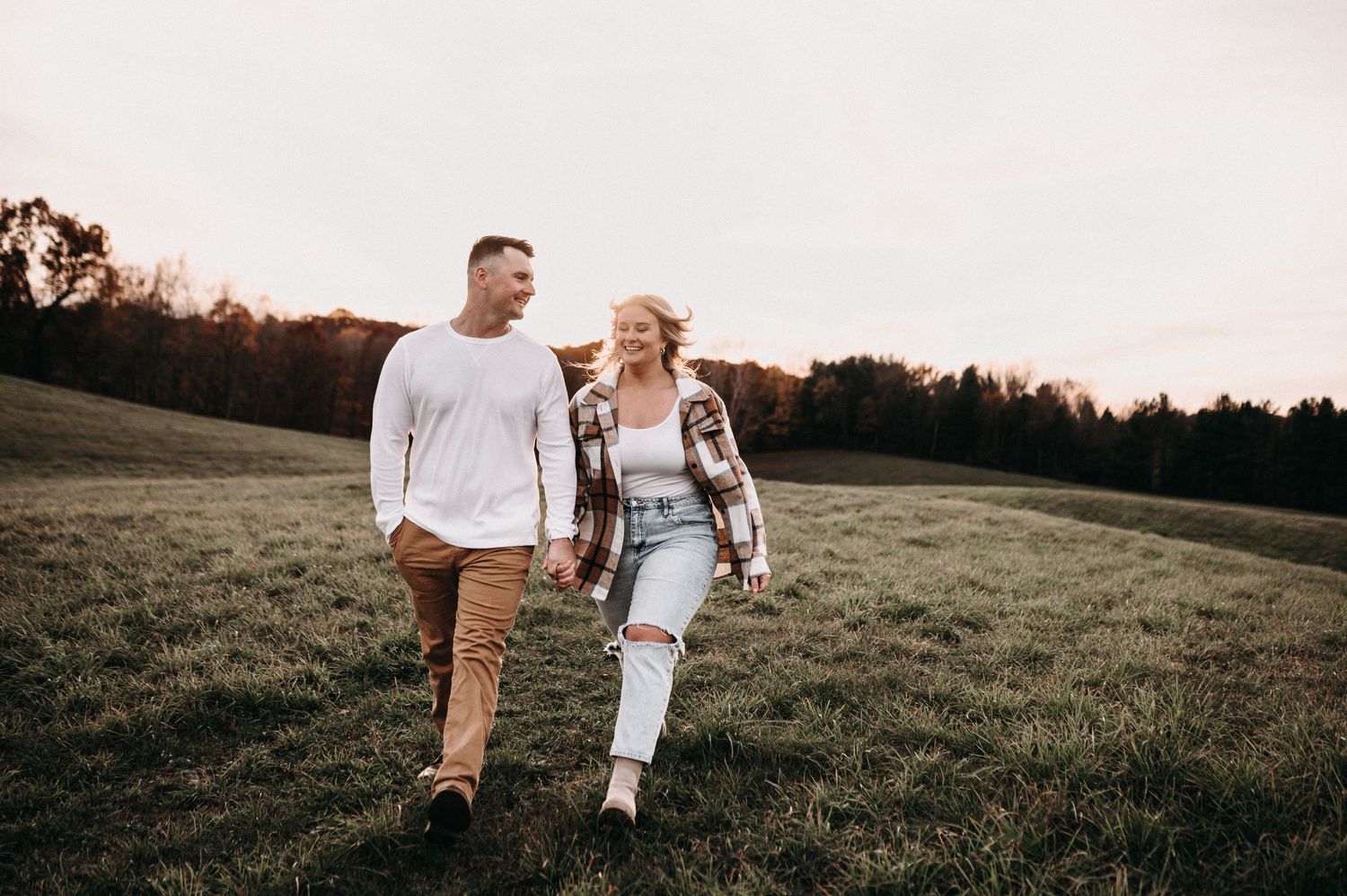 A couple walks hand in hand through a grassy field at sunset with trees in the background.