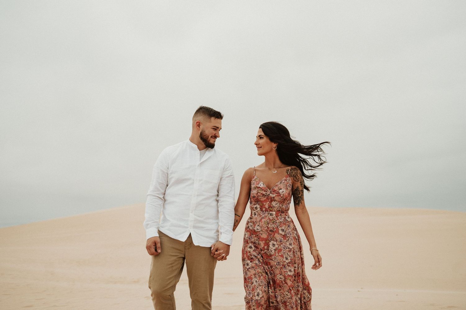 A couple in coordinated outfits walks hand in hand across desert sand dunes.