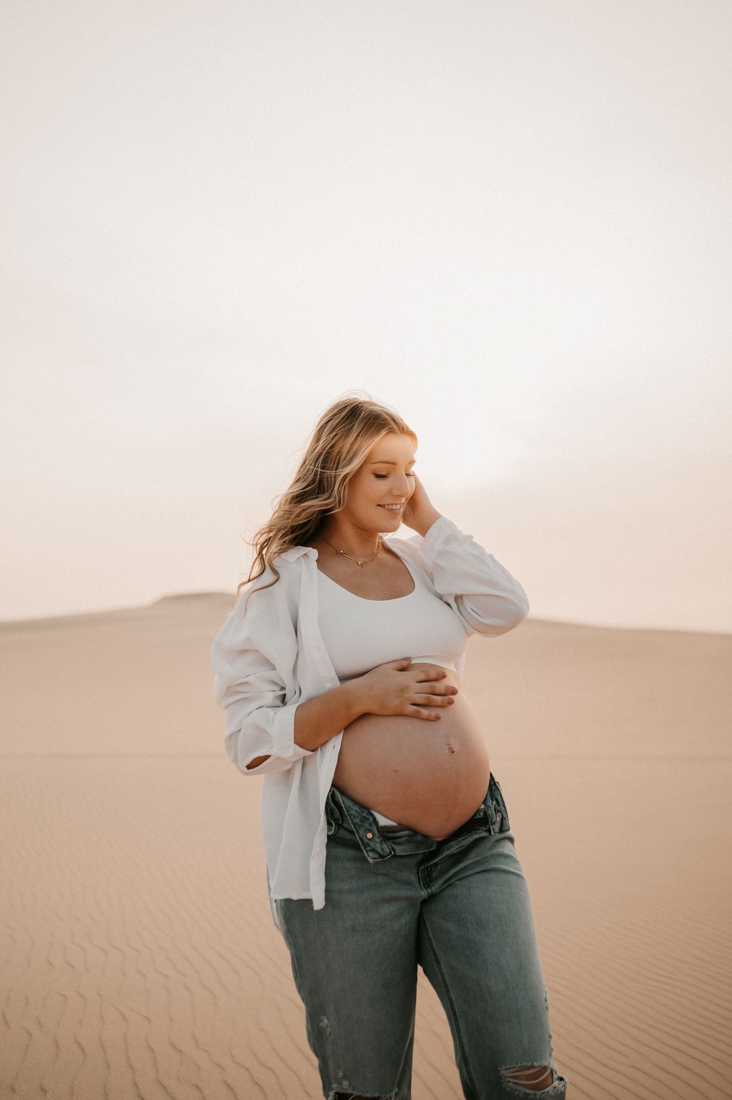 Expectant mother in white shirt and distressed jeans poses in desert setting.