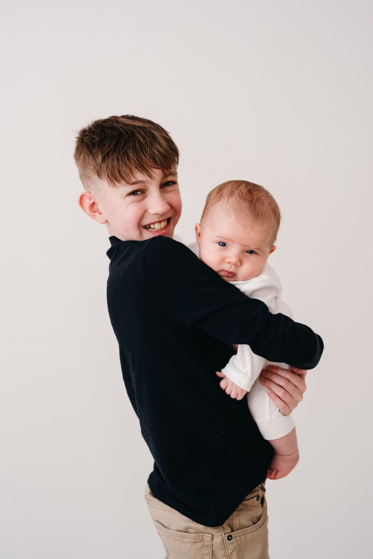 A young person in a black sweater holds and smiles while embracing a baby in white.