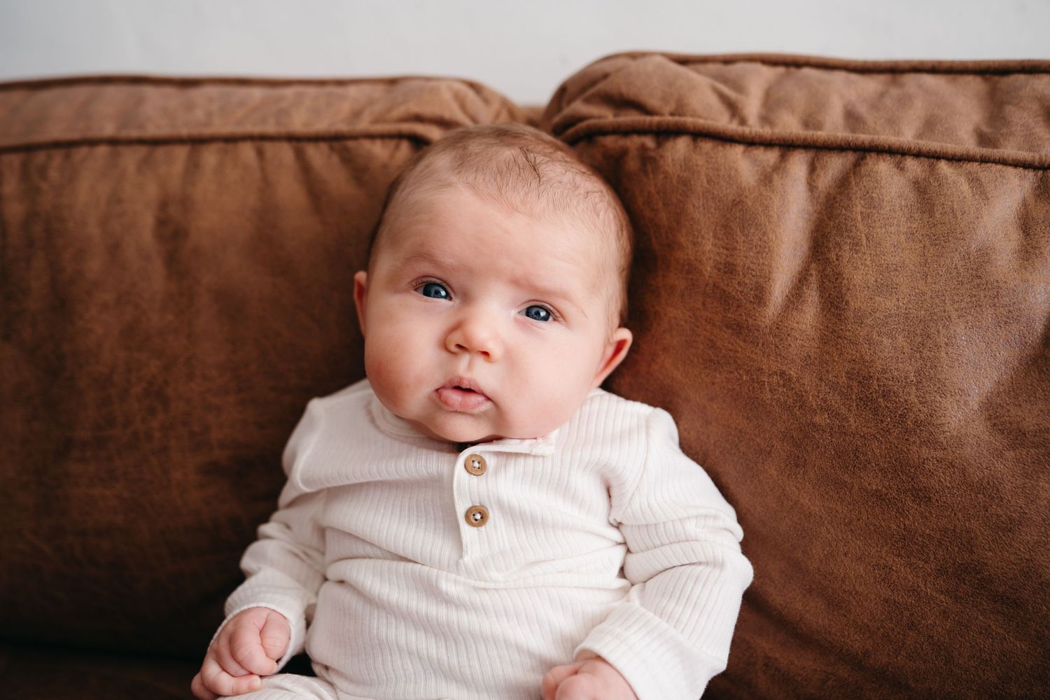 Baby wearing white buttoned sweater sits on brown leather couch looking at camera.