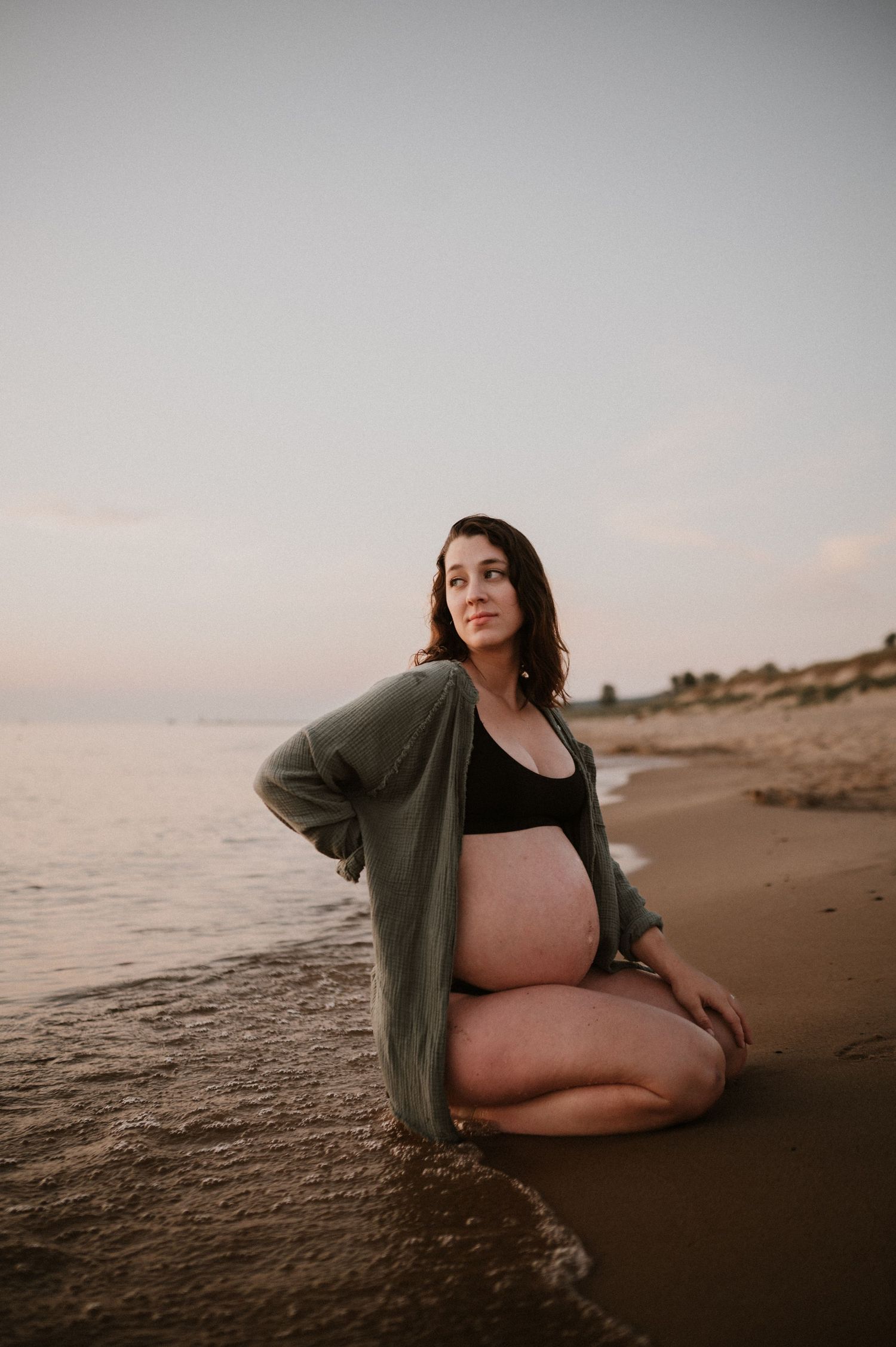 Expectant mother in black swimwear and gray cardigan poses on beach at sunset.