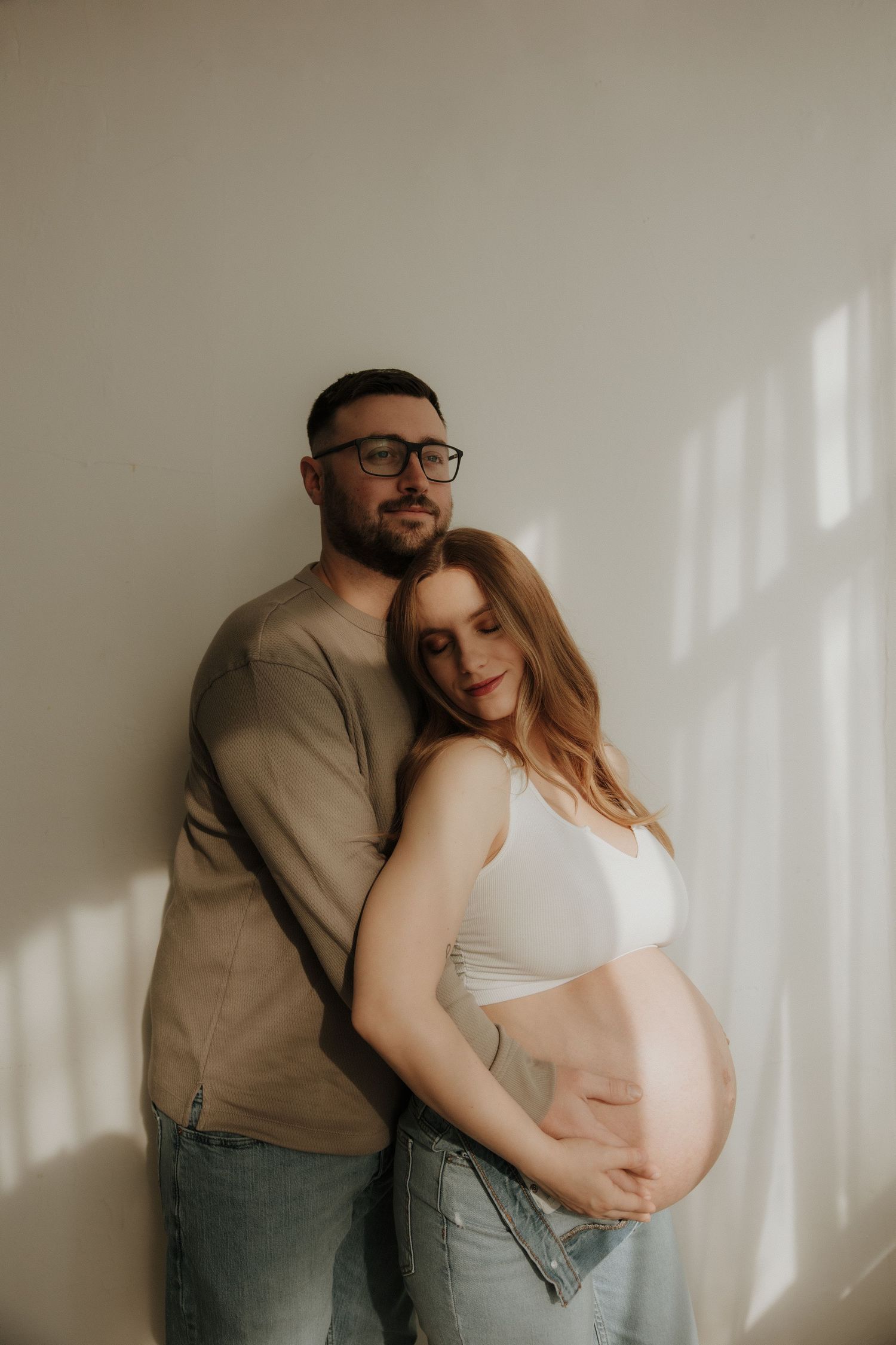 Intimate maternity photo of couple embracing against white wall with soft window light.