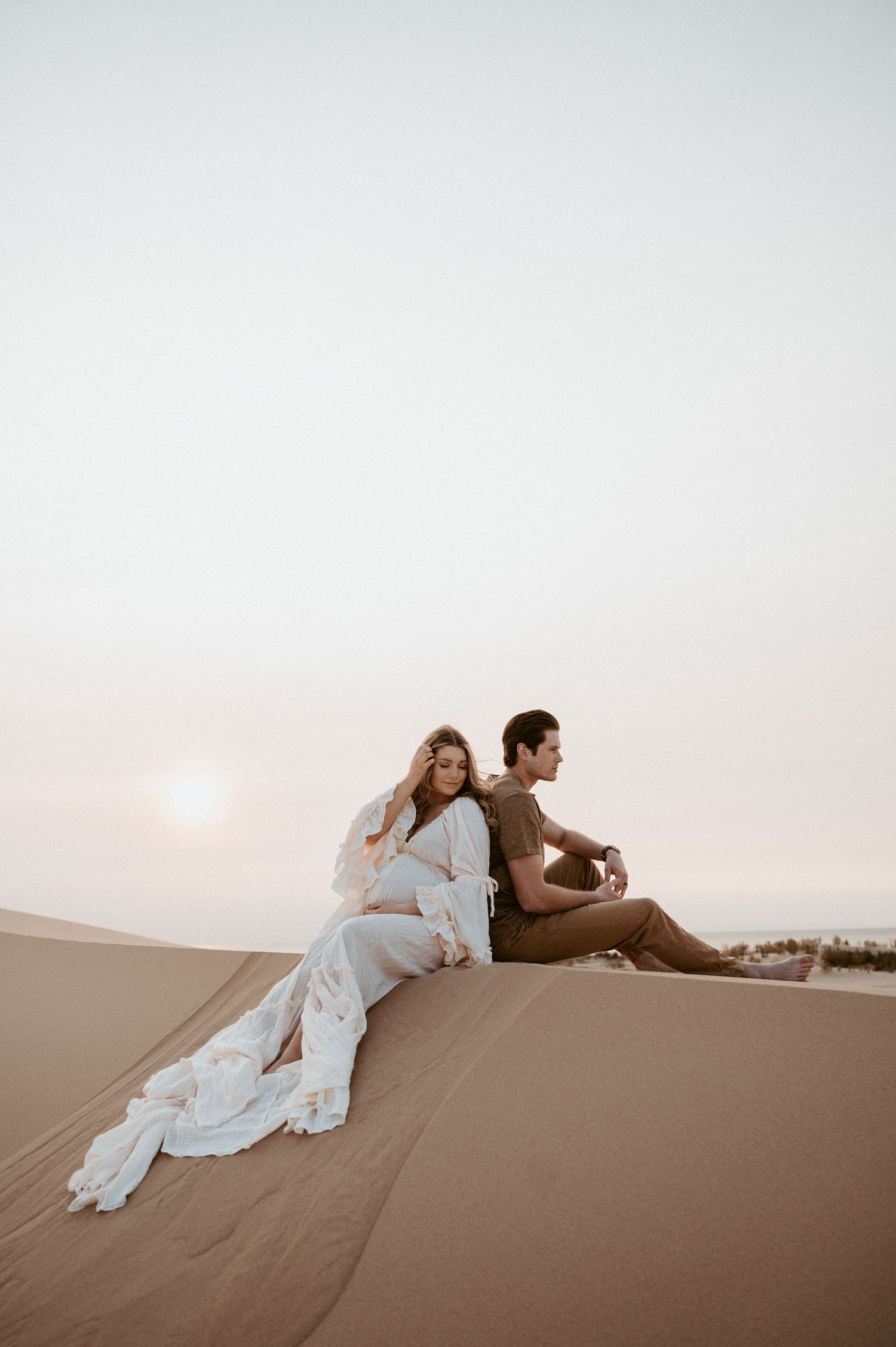Two people rest against each other on a desert dune during sunset.