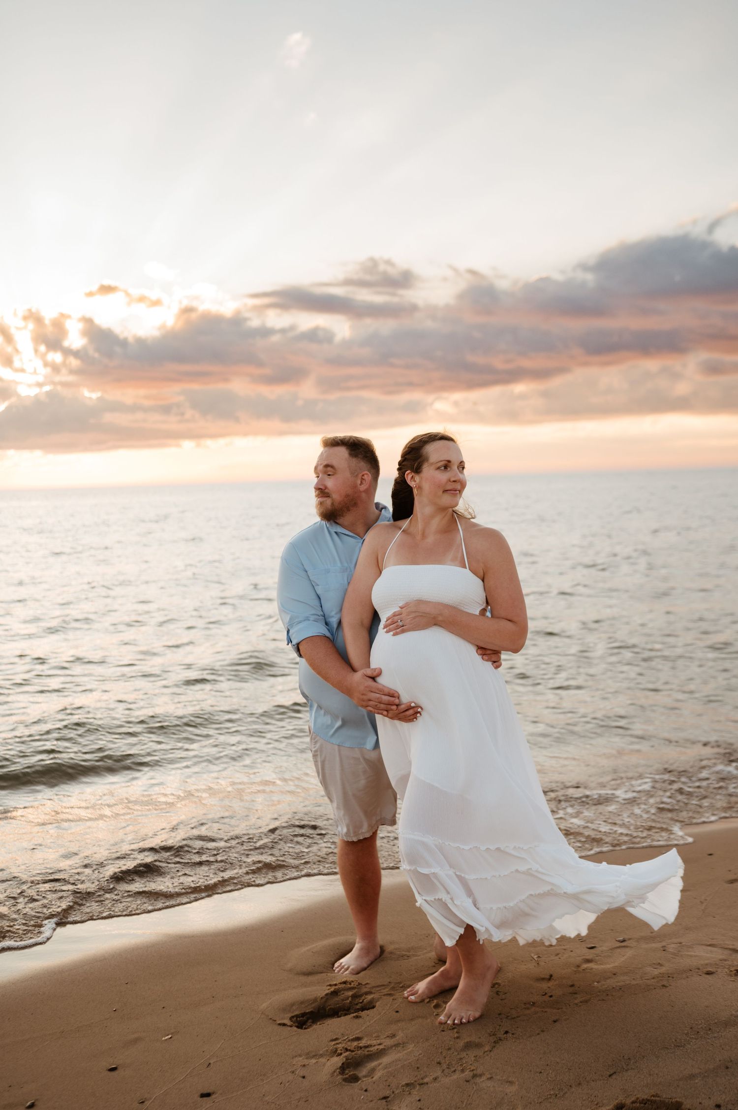 A couple embraces on a sandy beach at sunset while waves crash on the shore and orange clouds fill the sky.