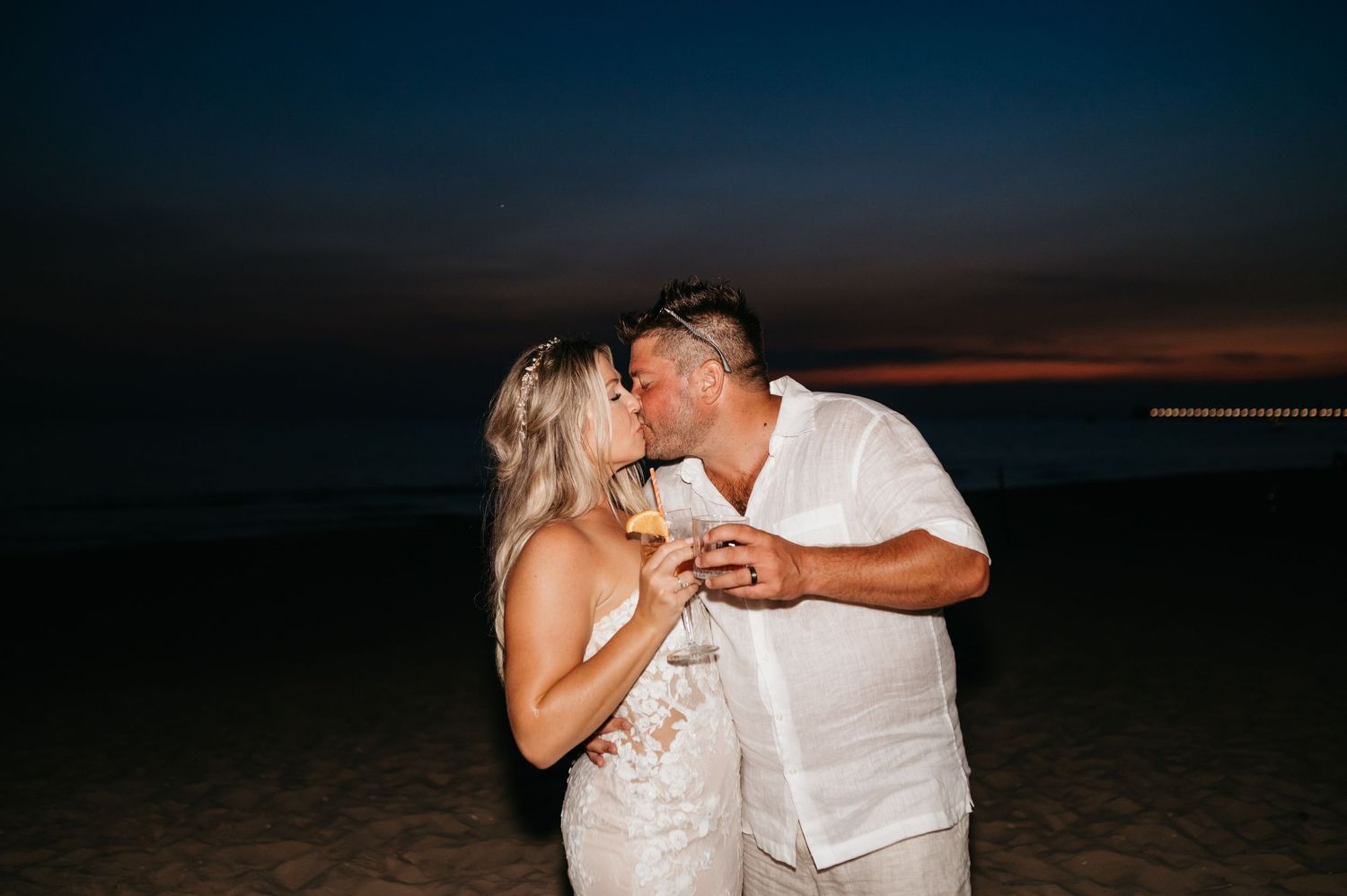 A couple shares a romantic moment on the beach at sunset while wearing casual white attire.