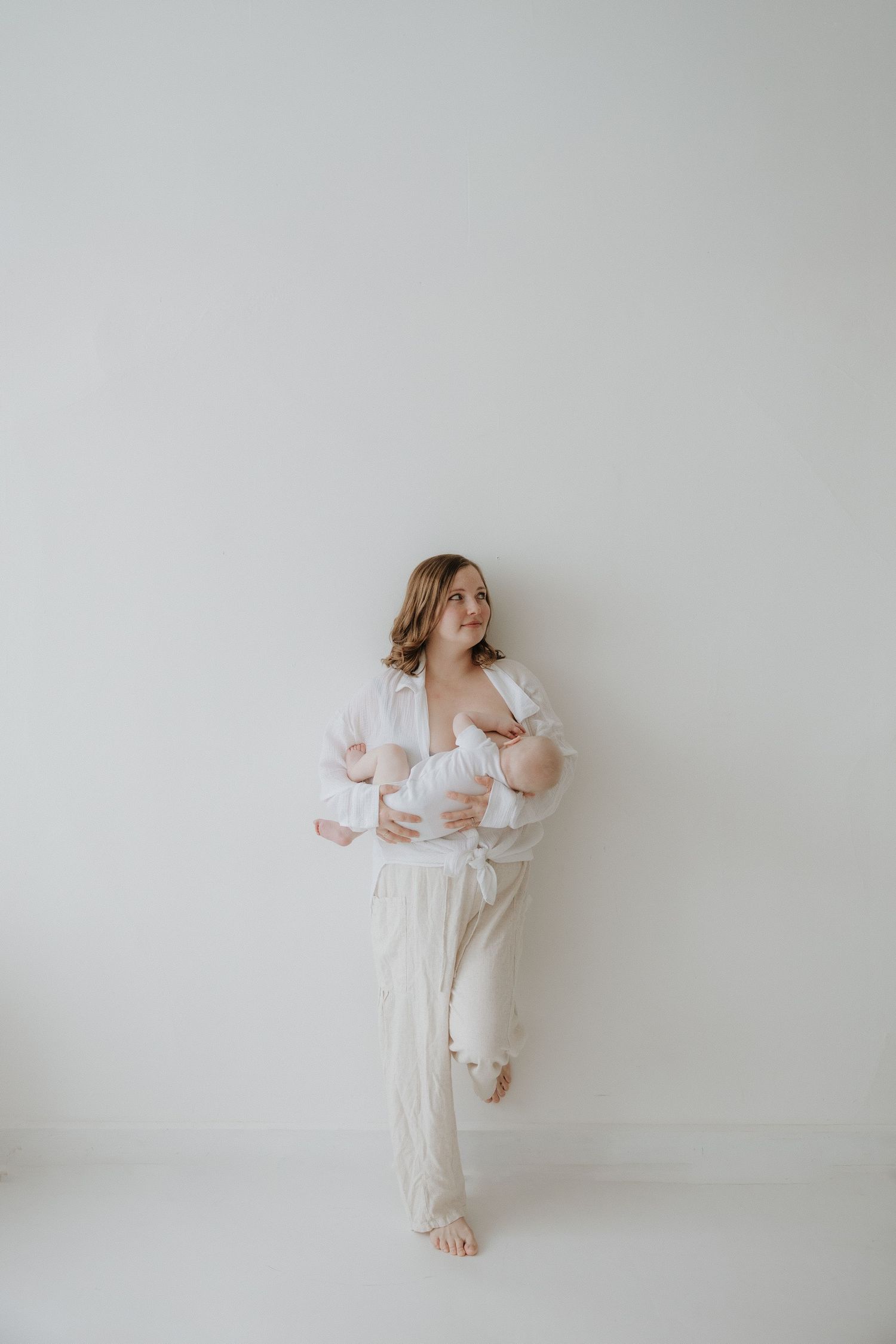 Mother in white outfit standing against white wall breastfeeding her newborn baby.