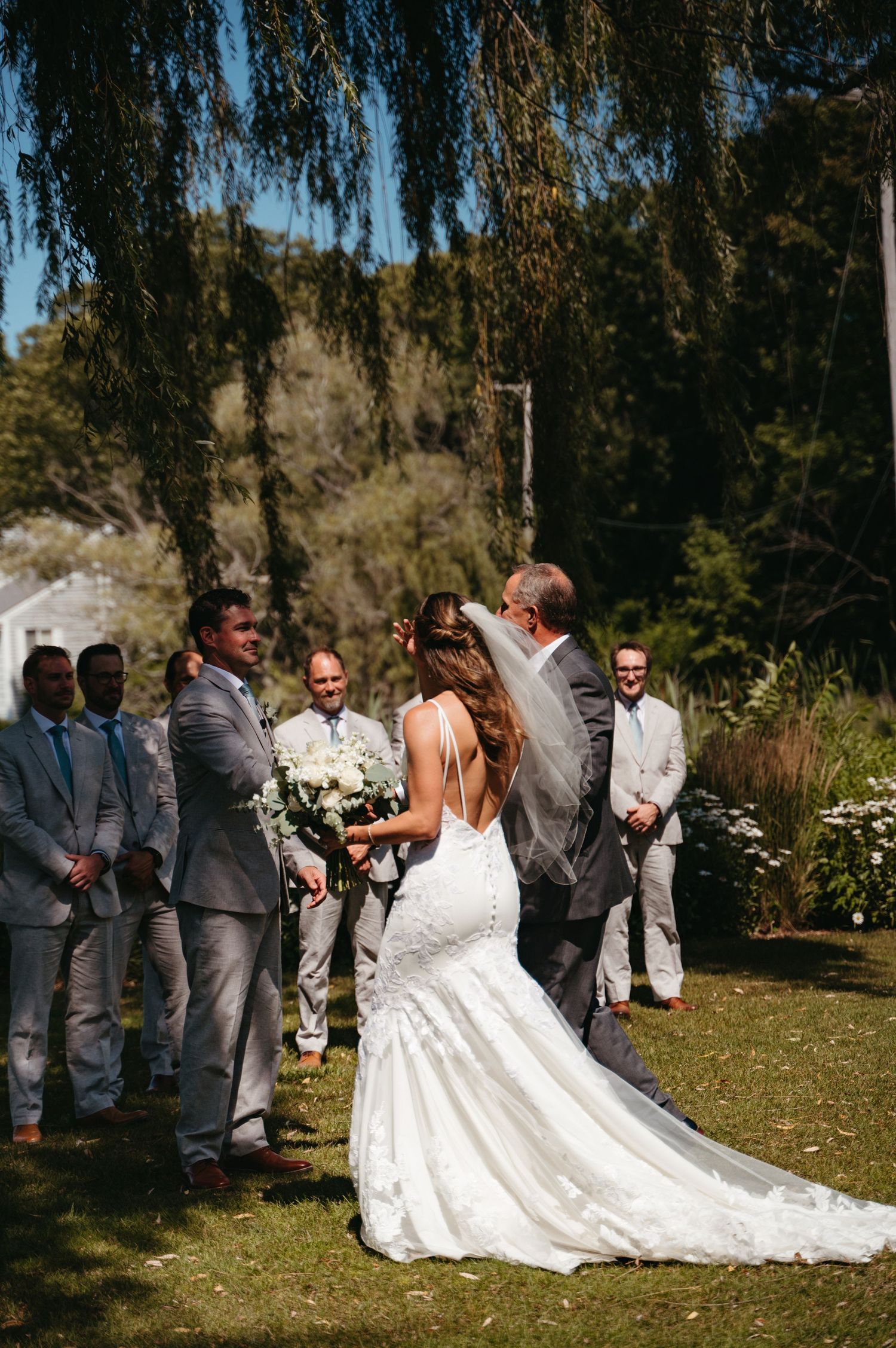 A wedding ceremony unfolds under a cascading willow tree with guests gathered on a grassy lawn.