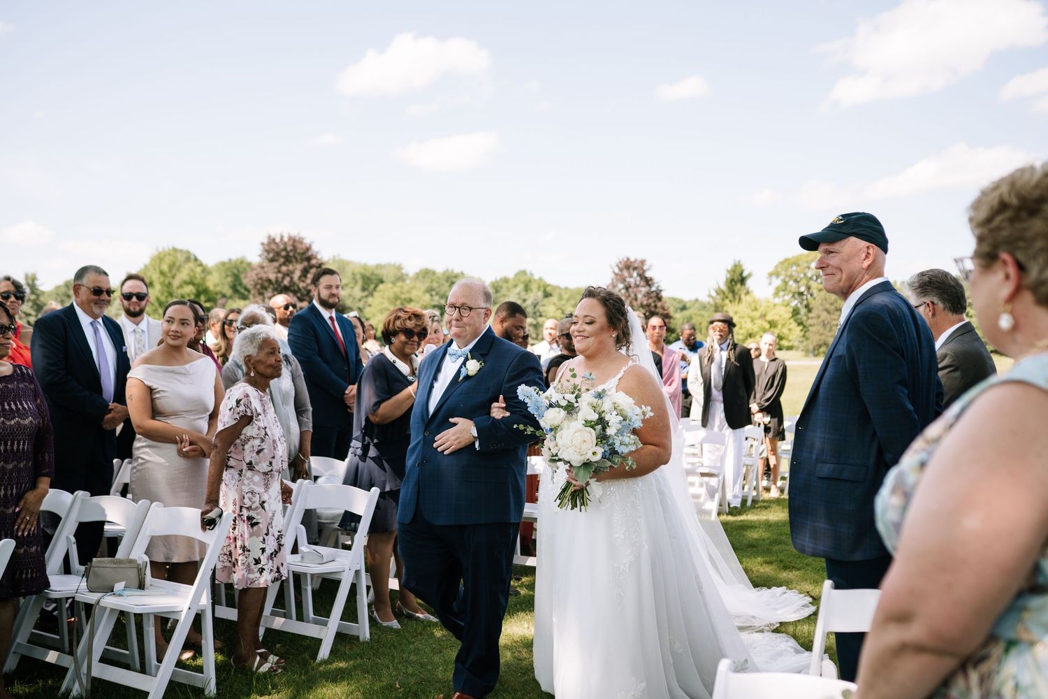 Outdoor wedding ceremony with guests seated in white chairs under blue sky.