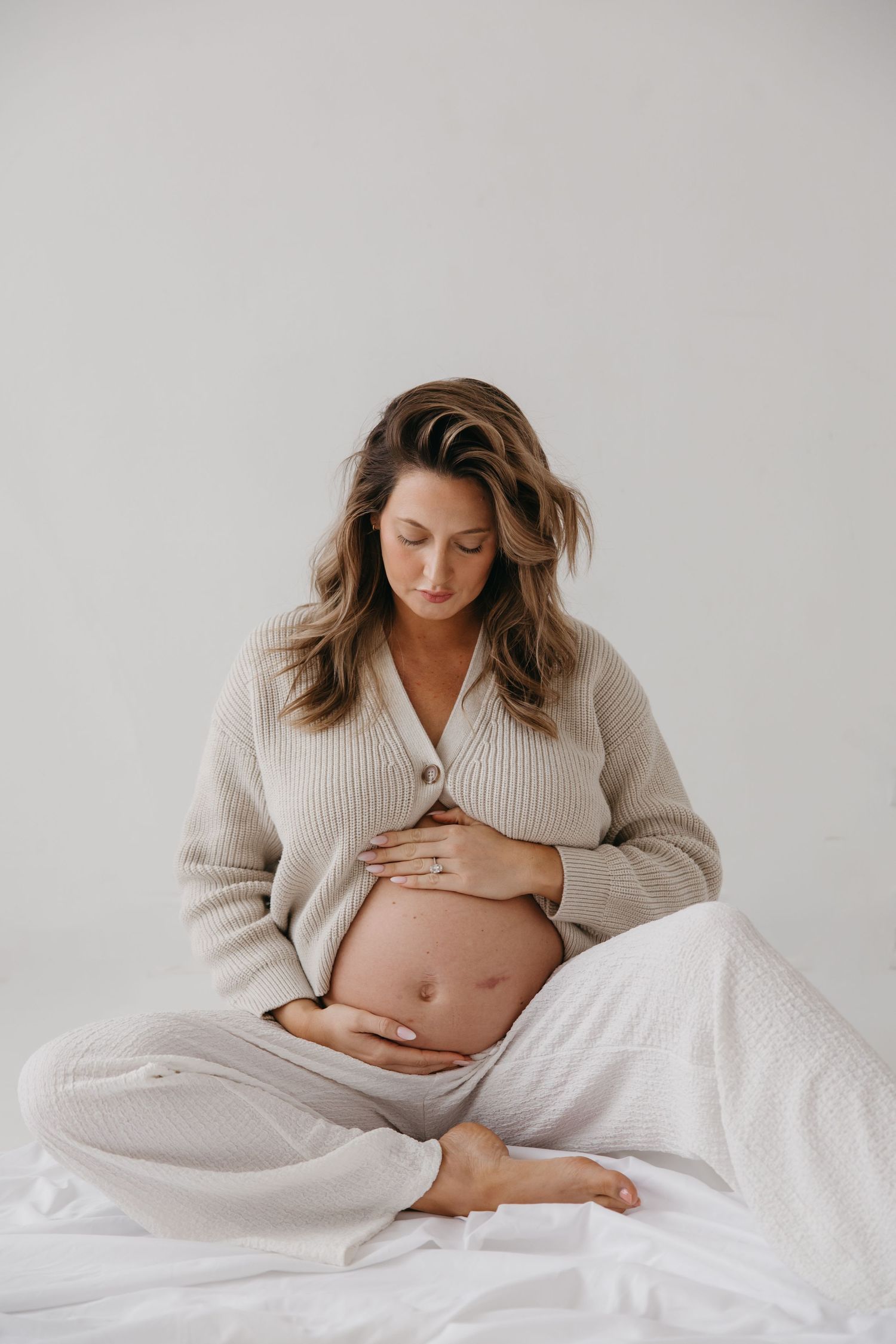 Pregnant woman in a beige ribbed knit cardigan sits cross-legged on white fabric, gently holding her bare baby bump.