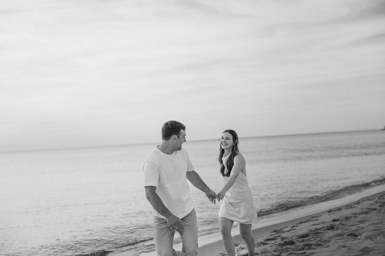 Black and white photo of a couple holding hands and walking along a serene beach shoreline.
