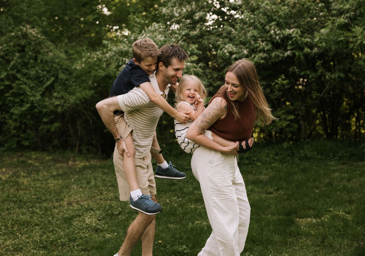 Happy family playing outdoors in lush green garden, parents carrying laughing children in warm summer setting.