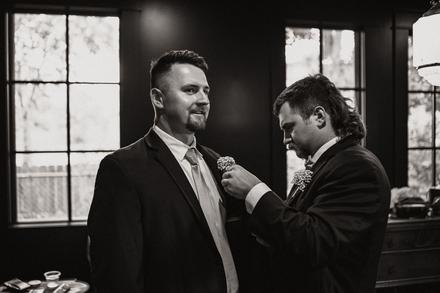Two people adjusting formal attire in a dimly lit room with window light.