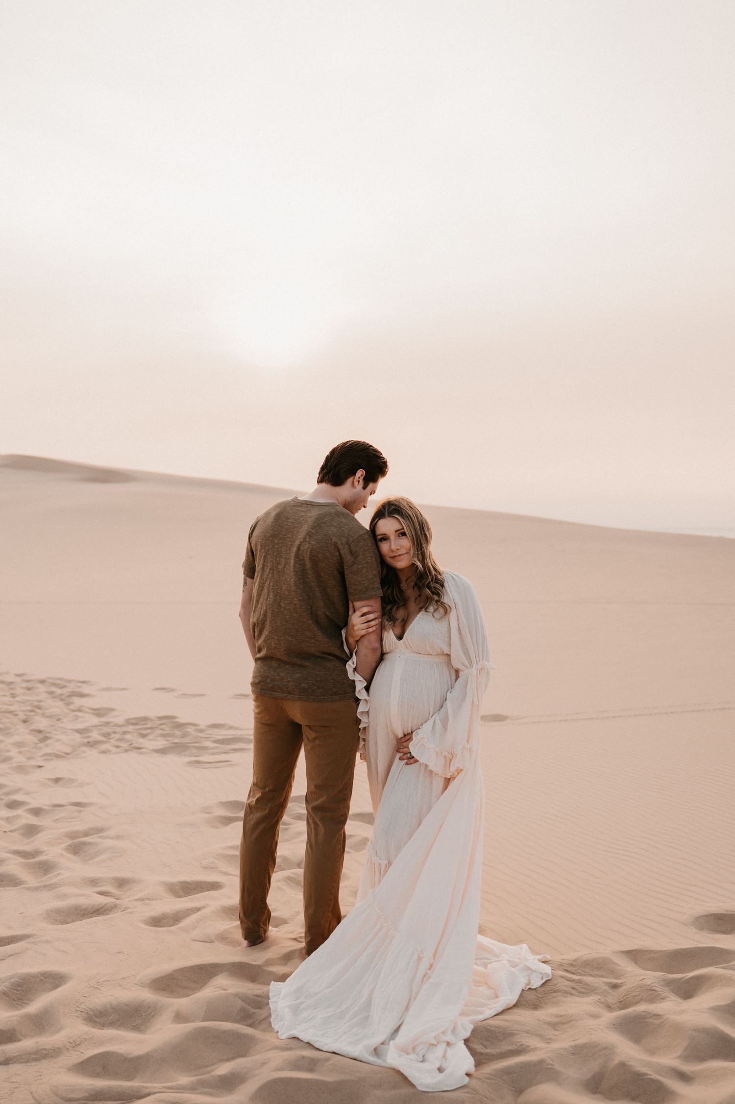Intimate desert maternity photo of couple in elegant attire at sunset.