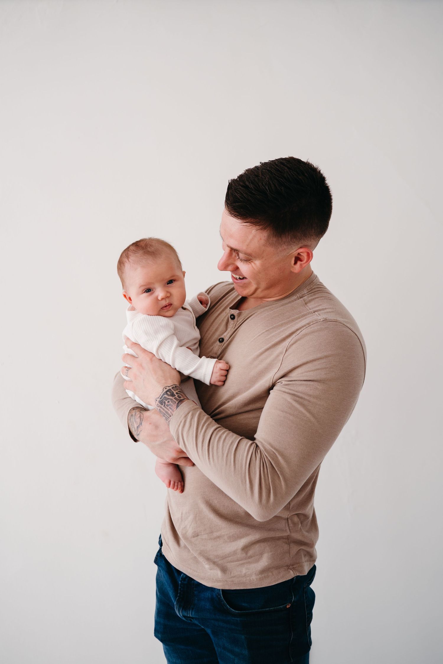 Parent in beige sweater holds and gazes lovingly at infant wearing white against neutral background.