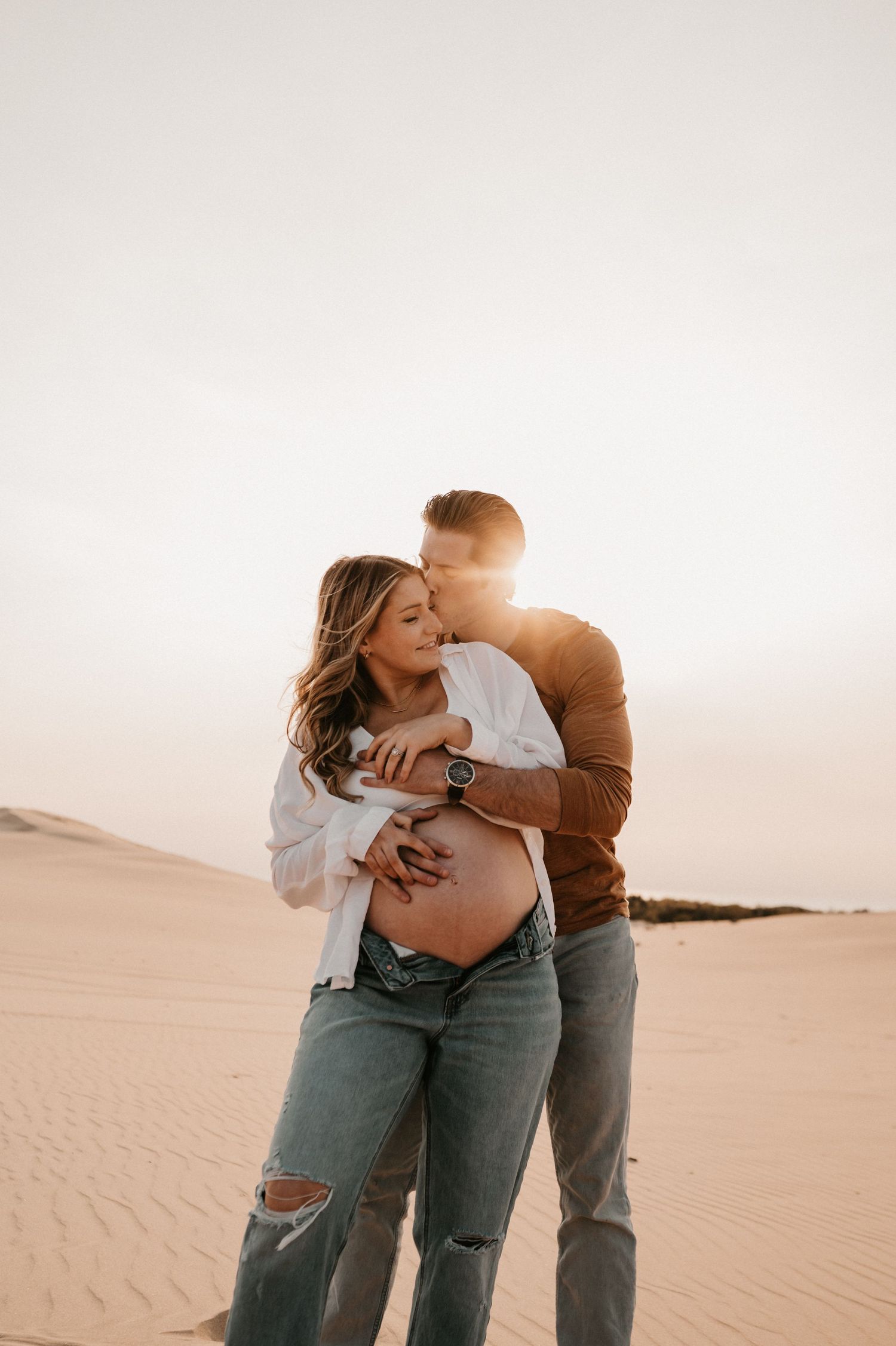 A tender maternity moment captured against desert sand dunes at sunset.