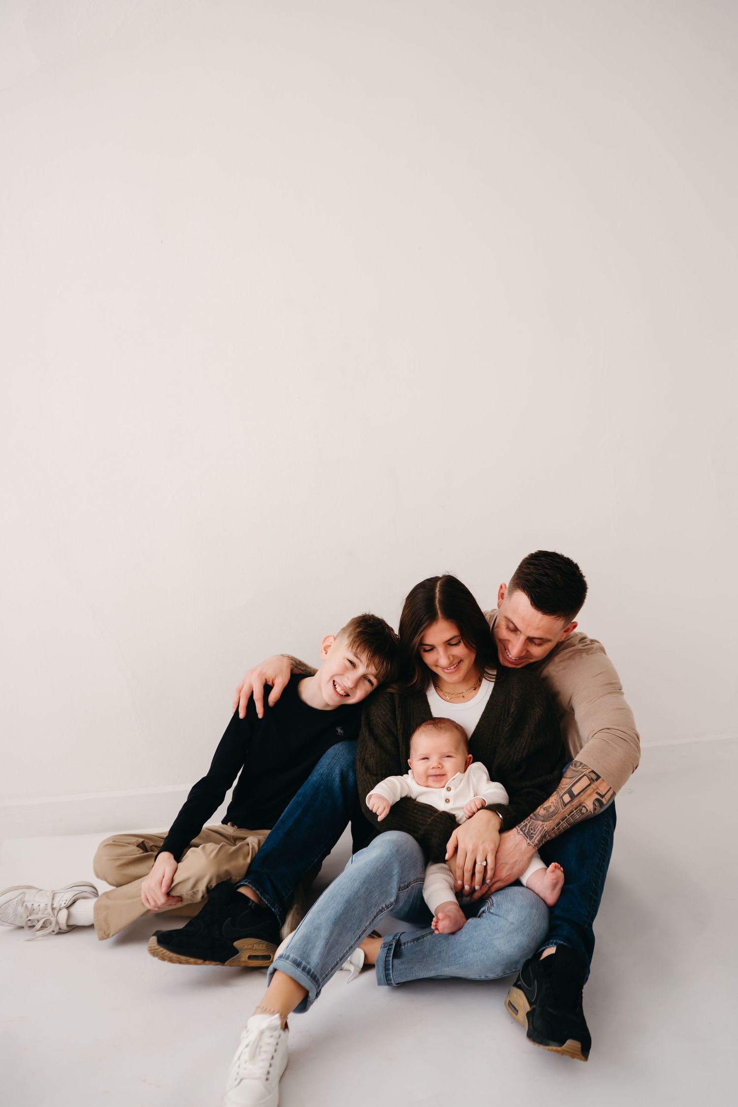 A family sits together on the floor against a white wall in a casual minimalist portrait style photo.