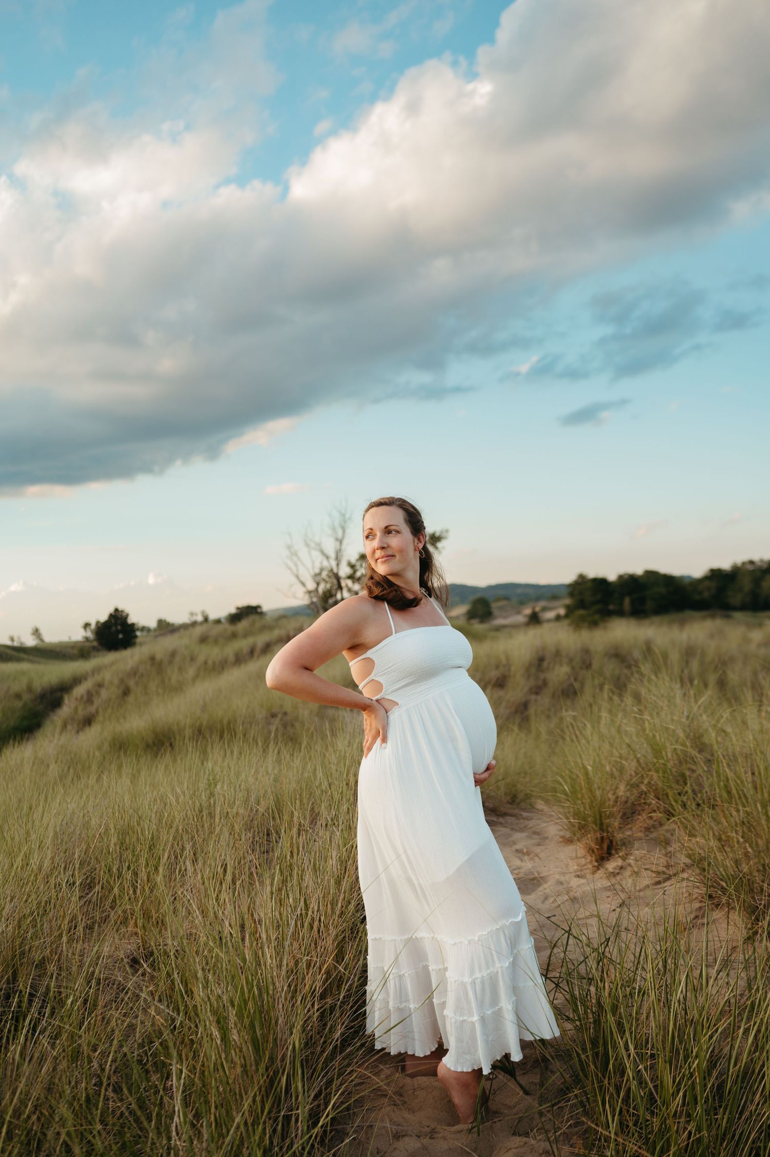 A person in a flowing white dress stands among tall grass on a hilltop during golden hour with cloudy skies above.