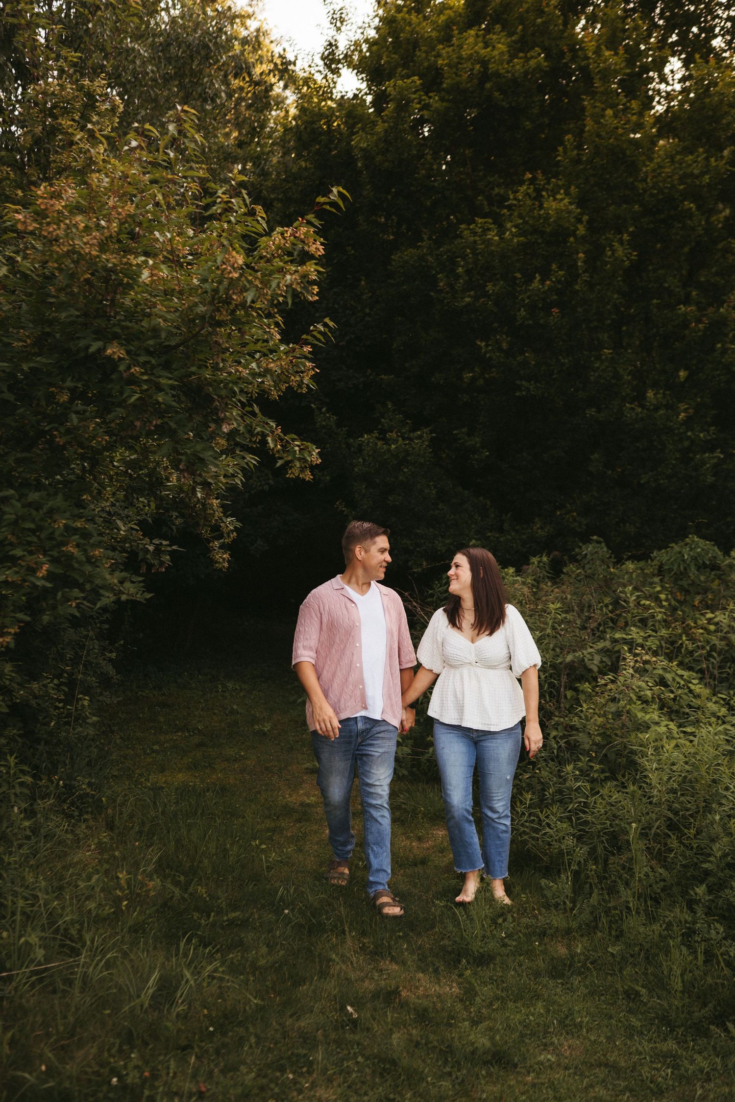 Couple in matching white tops and jeans walking together through a lush green garden at sunset.