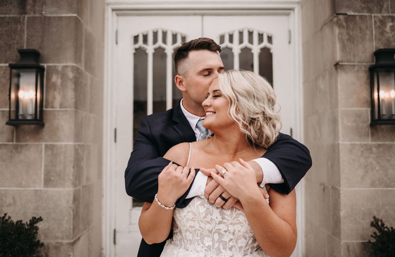 Intimate moment between newlyweds sharing laughter against stone church entrance.
