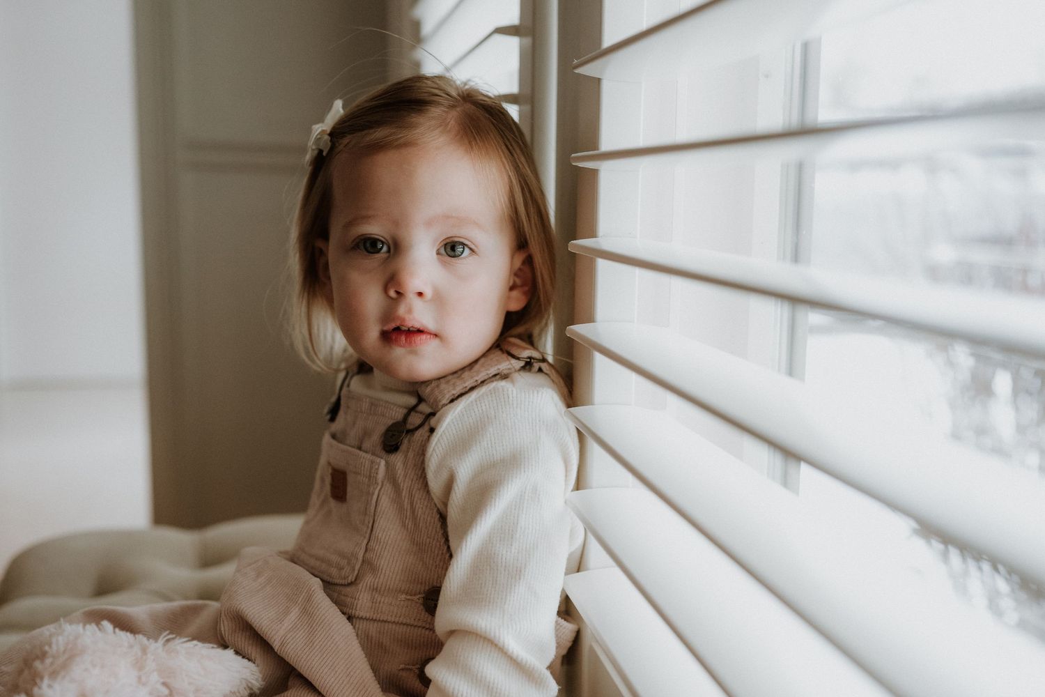 A young child in overalls and cream sweater sits thoughtfully by a window with blinds.